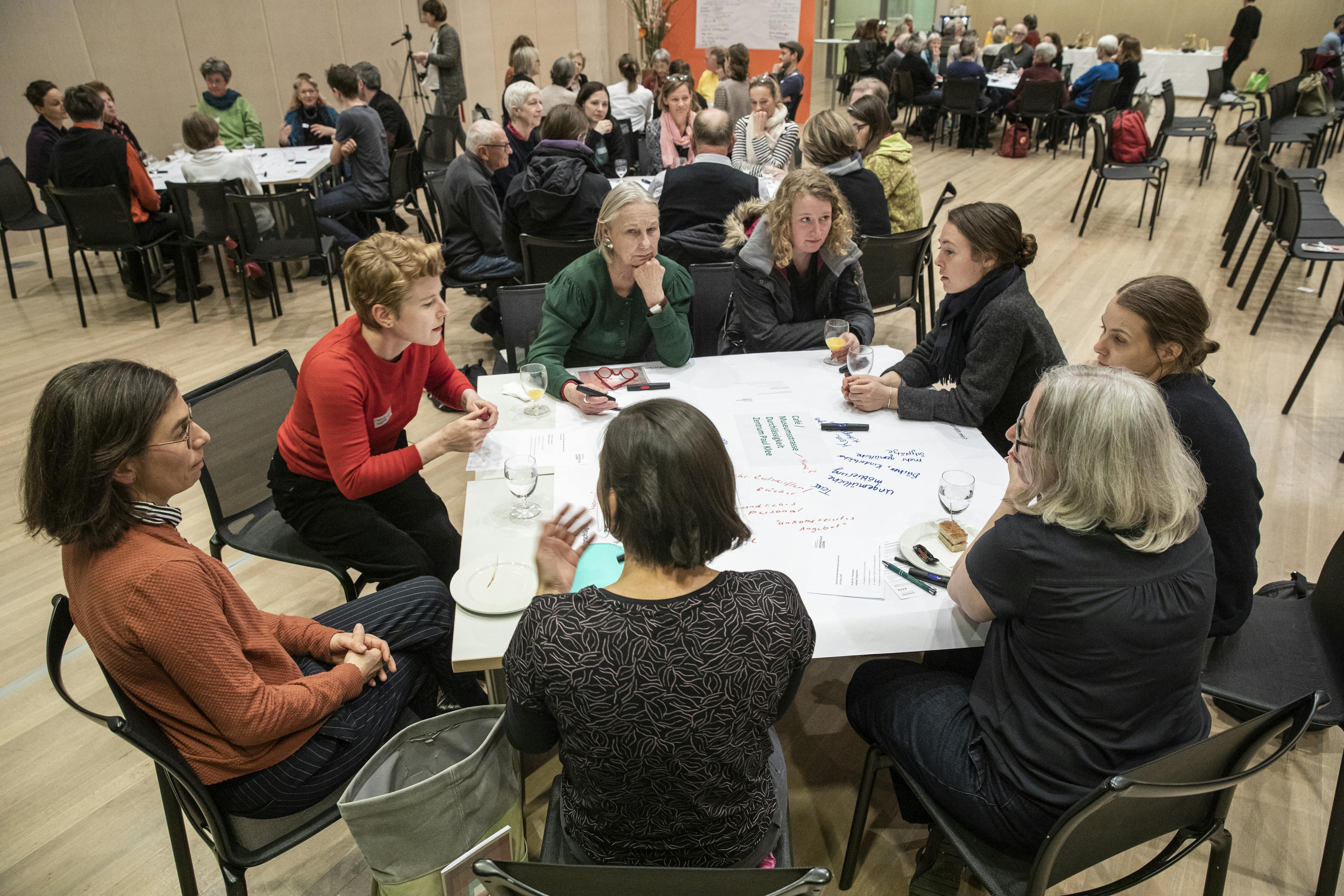 Eine Gruppe von Nachbar:innen und Mitarbeitenden des Zentrum Paul Klee sitzt um einen tisch und diskutiert Ideen. Im Hintergrund sind weitere Tischgruppen sichtbar.